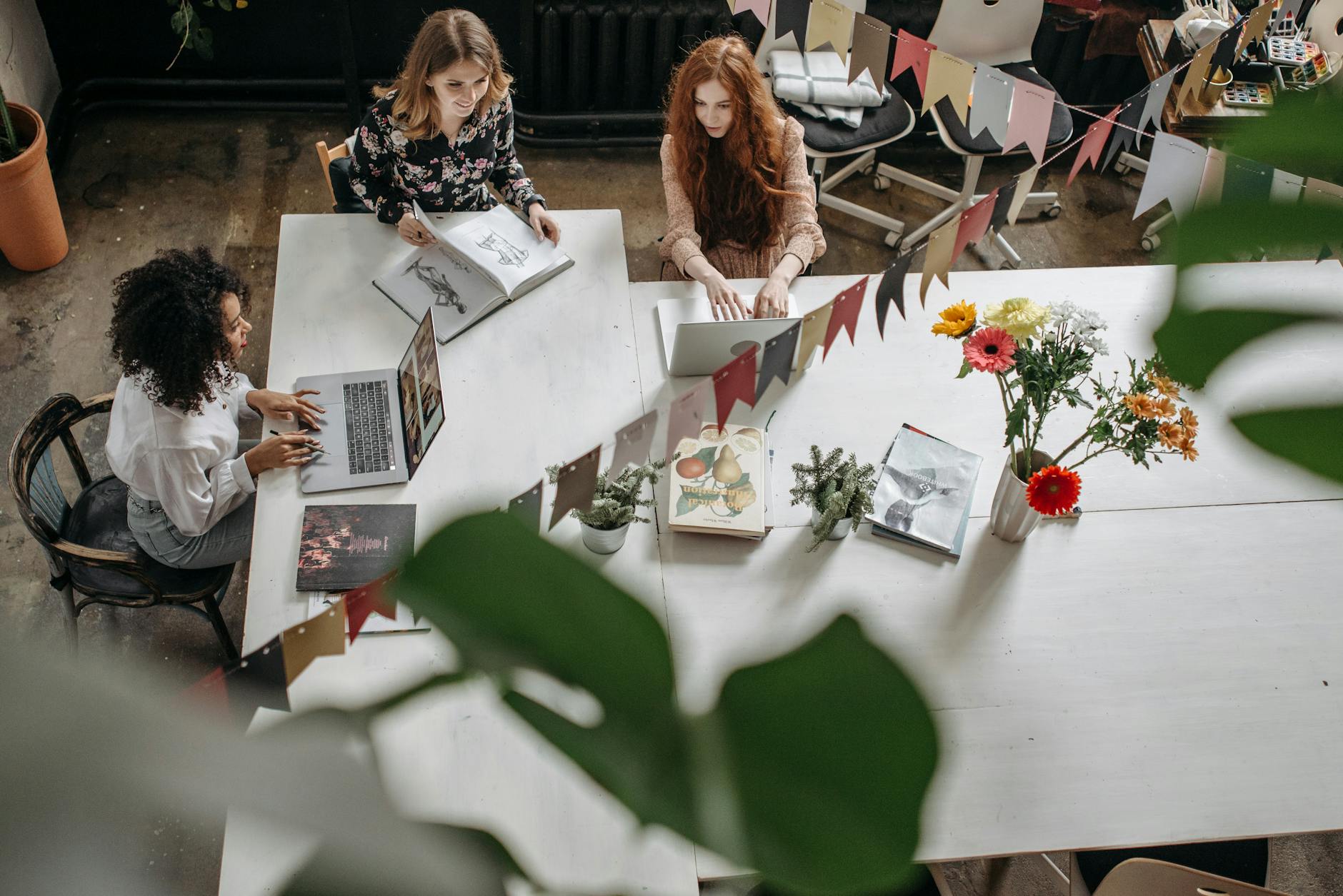 Women collaborating at a table with laptops, flowers, and books, representing a creative workspace.